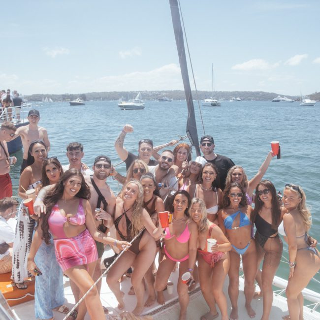A group of people in swimwear gathering and posing for a photo on a boat, with a body of water and other boats in the background. Some are holding drinks under the bright, sunny sky overhead, creating the perfect atmosphere for a catamaran party on Sydney Harbour.