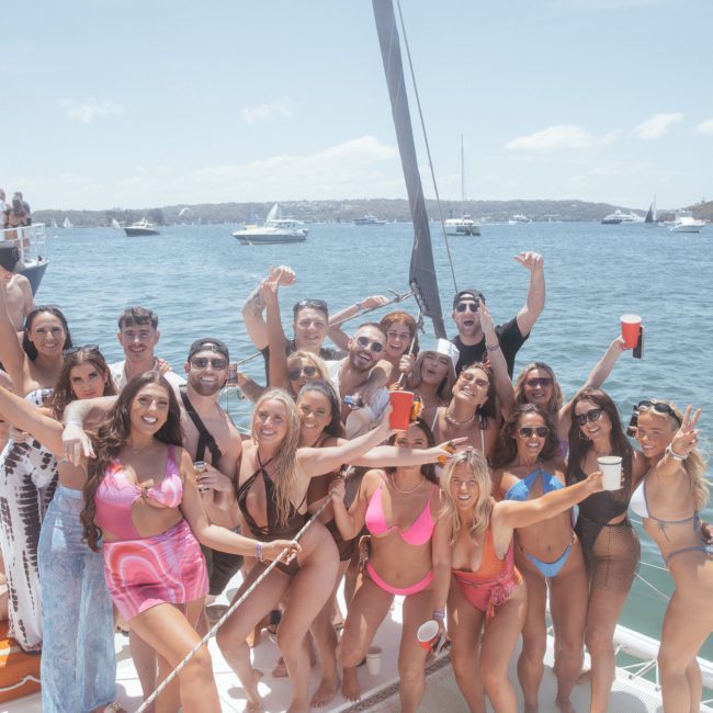 A group of people in swimwear are posing for a photo on a boat, with some holding drinks. Other boats and a shoreline are visible in the background—perfect for a Sydney boat party hire.
