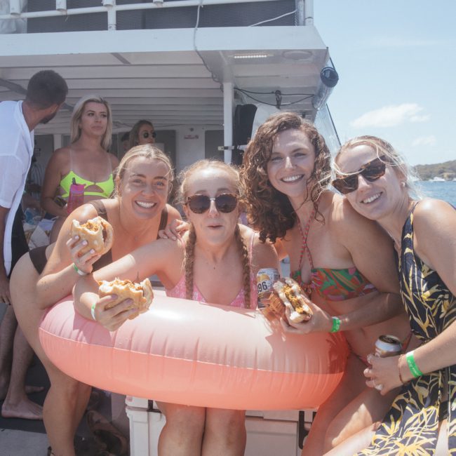 A group of people on a boat, with four women in the foreground smiling and holding burgers, one wearing an inflatable ring, enjoying a sunny day by the water. Perfect for a Sydney boat party hire!