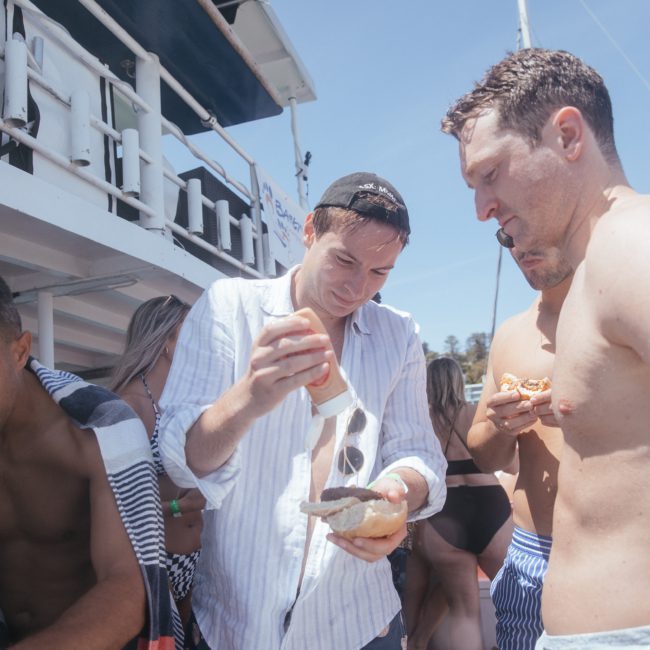 A man in a white shirt and patterned shorts applies sauce to a piece of bread, surrounded by people in swimwear on a luxurious catamaran party in Sydney.