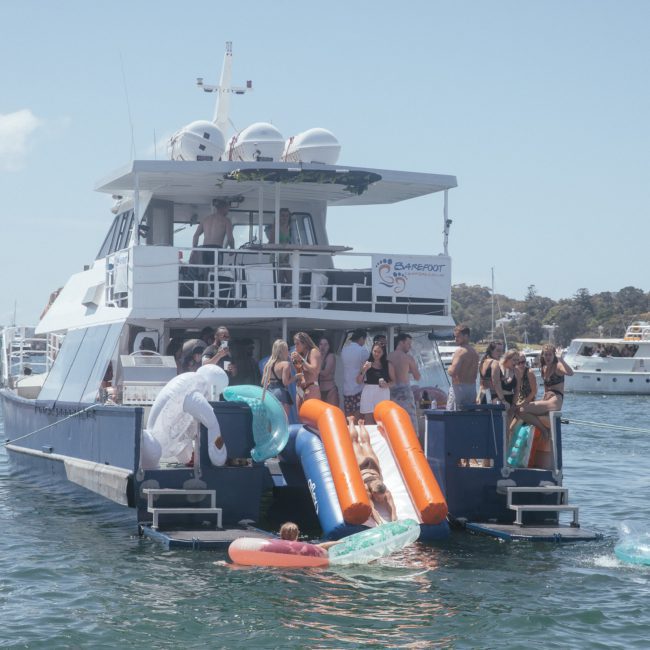 People enjoy water activities from the back of a moored boat in a scenic area with several other boats in the background, perfect for Sydney boat party hire.