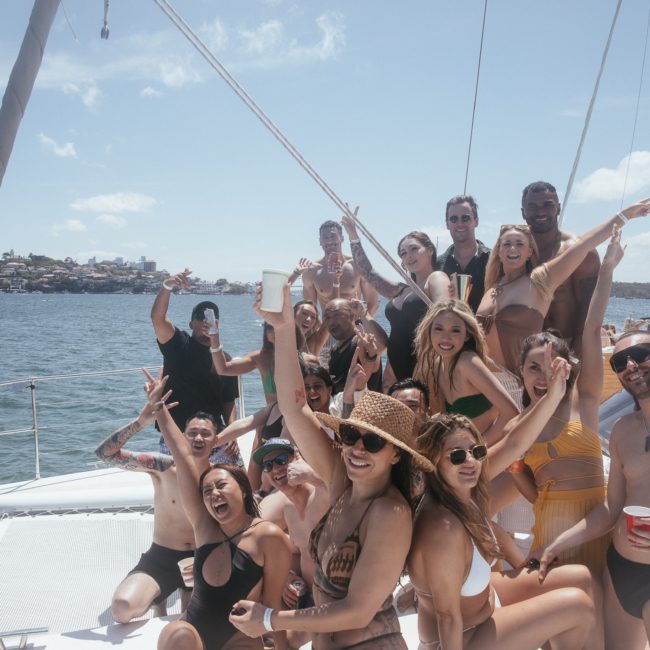 A large group of people enjoying a sunny day on a catamaran party in Sydney, some sitting and others standing, with smiles and raised arms, the ocean and distant shoreline visible in the background.