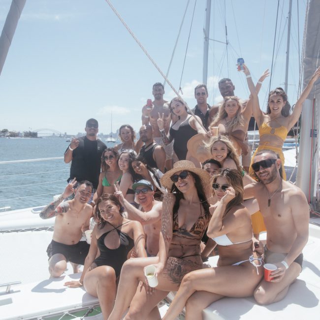 A group of people in swimsuits poses for a photo on a sunny day aboard a private yacht charter in Sydney Harbour. They are smiling, with some holding drinks. The ocean and a city skyline are visible in the background.