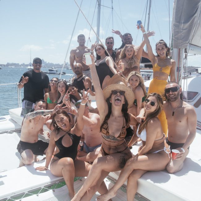 A group of people posed on a catamaran party in Sydney, smiling and holding drinks, with a sunny sky and water in the background.