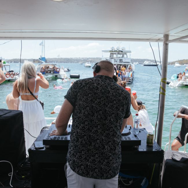 A DJ plays music on a luxury yacht hire Sydney while people in swimsuits dance and swim in the surrounding water.