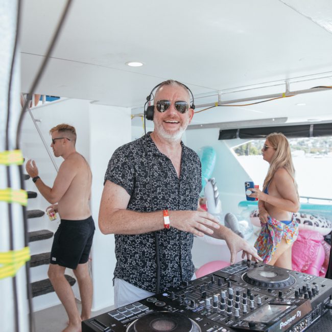 A person wearing sunglasses and a patterned shirt is DJing on a private yacht charter in Sydney Harbour. Other individuals are seen socializing in the background in swimwear.