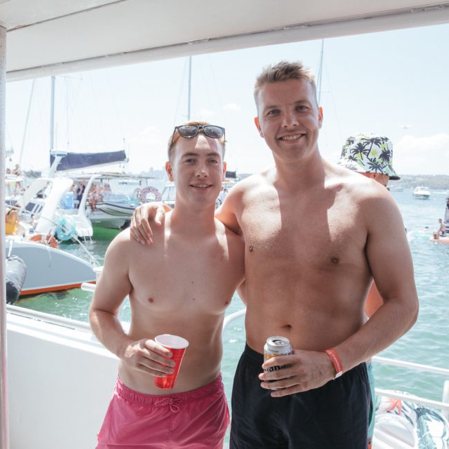 Two men in swim trunks smile and pose for a photo on a boat. One holds a red cup, and the other holds a can. Boats and people are visible in the background on a sunny day, giving off the vibe of an exciting Sydney boat party hire.