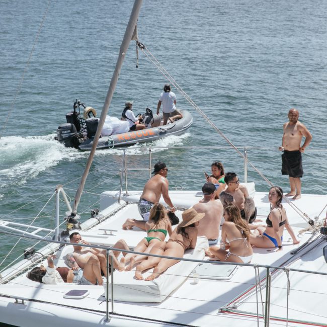 A group of people are sunbathing and socializing on a luxury yacht hire in Sydney Harbour, while another boat with people passes by on the water. The background includes a shoreline with buildings and trees.