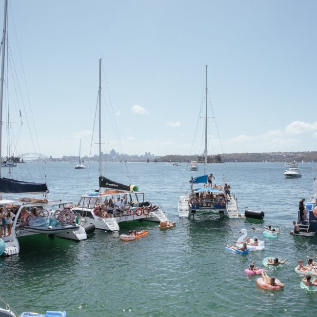 A group of boats and yachts anchored close together in a sunny bay, with people enjoying the water on floating inflatables and slides. Sydney boat party hire offers an unforgettable experience with the city skyline visible in the distance.