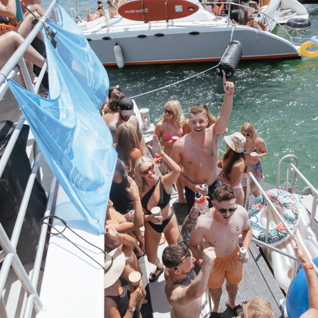 A group of people in swimsuits are gathered on a luxury yacht hire in Sydney, holding drinks and celebrating. Other boats and inflatable rings are visible in the water, making it the perfect scene for a memorable Sydney boat party.