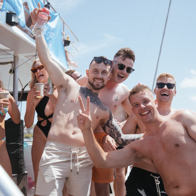 A group of people in swimsuits are smiling and posing for a photo on a private yacht charter Sydney Harbour under clear skies, some holding drinks.