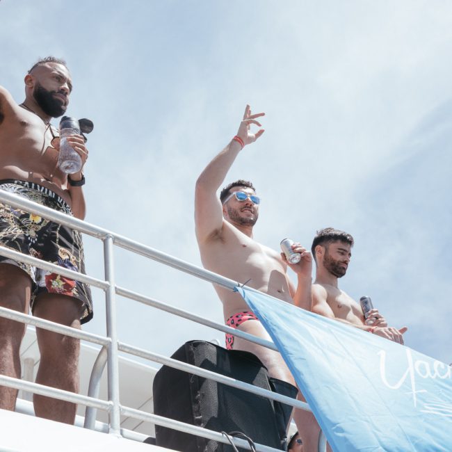 Three men in swim trunks are standing on a boat deck, holding drinks and gesturing. There is a blue flag hanging below them. The sky is clear and blue, perfect for luxury yacht hire Sydney.