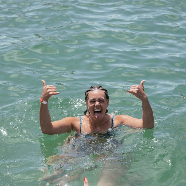 Person swimming in clear green water, smiling, and making a "hang loose" gesture with both hands near a luxurious yacht hire in Sydney.