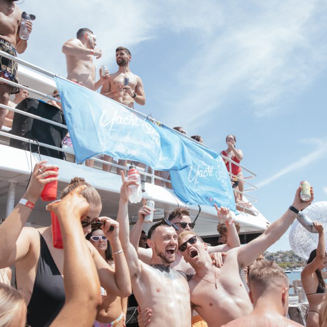 A group of people celebrating on a luxury yacht hire in Sydney, holding drinks and cheering. Blue banners with white text are hanging on the railing. The sky is clear and sunny.
