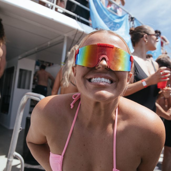 Person wearing reflective sunglasses and a pink bikini smiles widely at the camera on a private yacht charter in Sydney Harbour, surrounded by people under a blue sky.