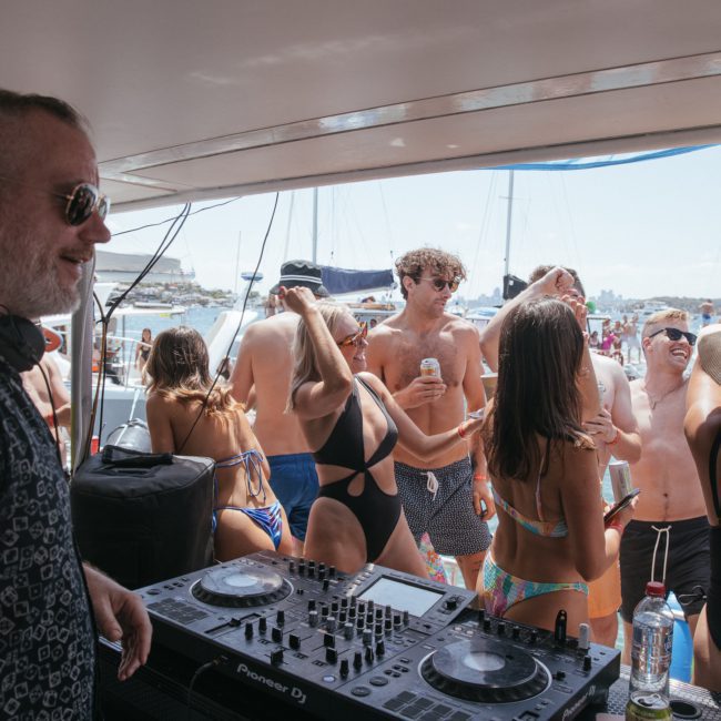 A DJ playing music on a private yacht charter in Sydney Harbour while a crowd of people in swimwear dance and socialize under a sunny sky.