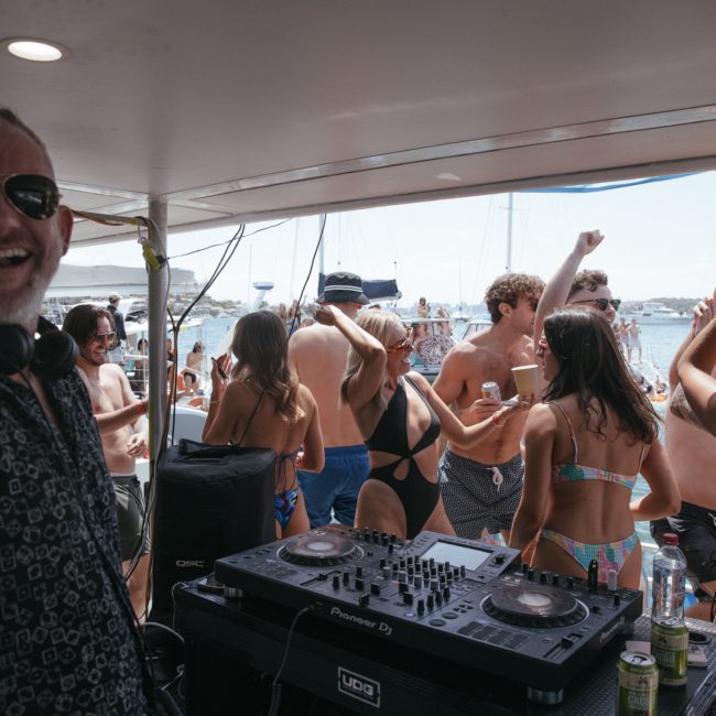 A man in sunglasses stands near DJ equipment on a boat, with a crowd of people in swimsuits dancing and socializing in the background. The scene overlooks the water and several boats, showcasing the vibrant atmosphere of a luxury yacht hire Sydney event.