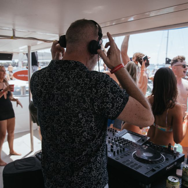 DJ with headphones performs on a private yacht charter Sydney Harbour while a crowd of people in swimwear dances and mingles in the background.