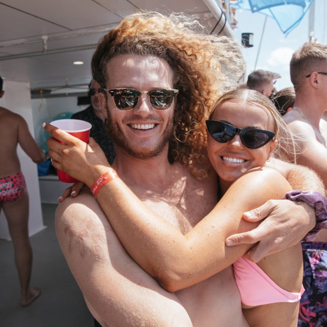 A man and woman in swimsuits, both wearing sunglasses, smile while hugging on a boat crowded with other people. The man holds a red cup on a sunny day during their private yacht charter Sydney Harbour experience.