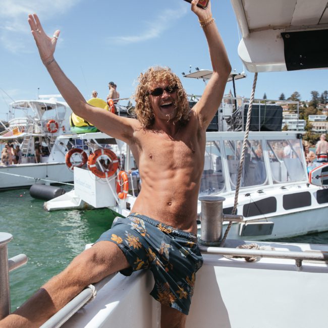 A shirtless man with curly hair and sunglasses poses with raised arms on a boat, with multiple boats and people in the background on a sunny day, embodying the fun atmosphere of a catamaran party in Sydney.