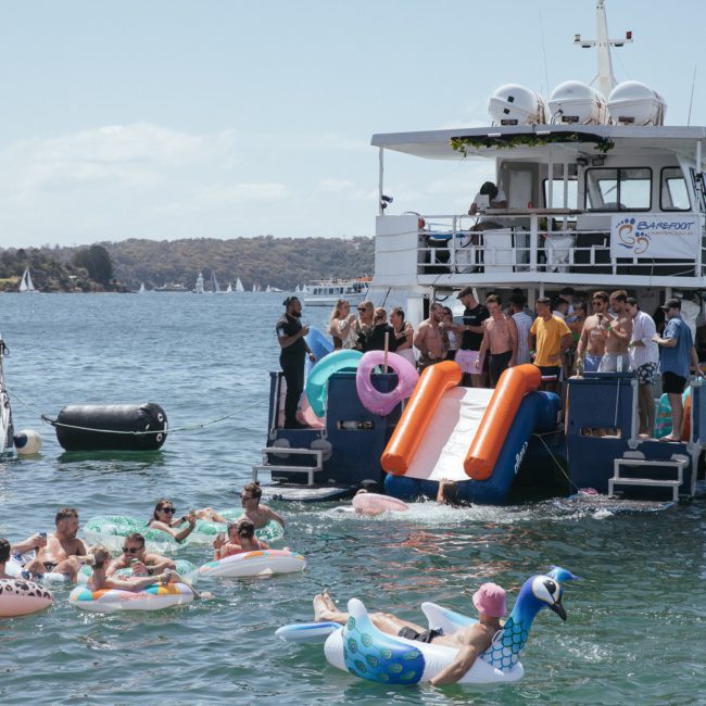 A group of people enjoying a sunny day on a luxury yacht hire in Sydney, with inflatable slides into the water, and others in the water floating on various inflatable toys.