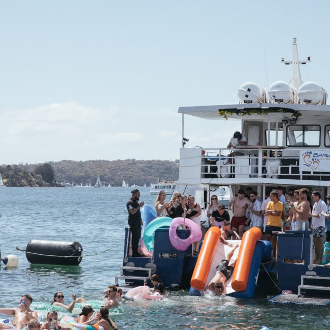 People are gathered on a boat with inflatables, enjoying a lively swim as they jump into the water. Other smaller boats and swimmers are nearby. The backdrop features distant hills and a partly cloudy sky, perfect for a Sydney boat party hire or DJ boat hire Sydney event.