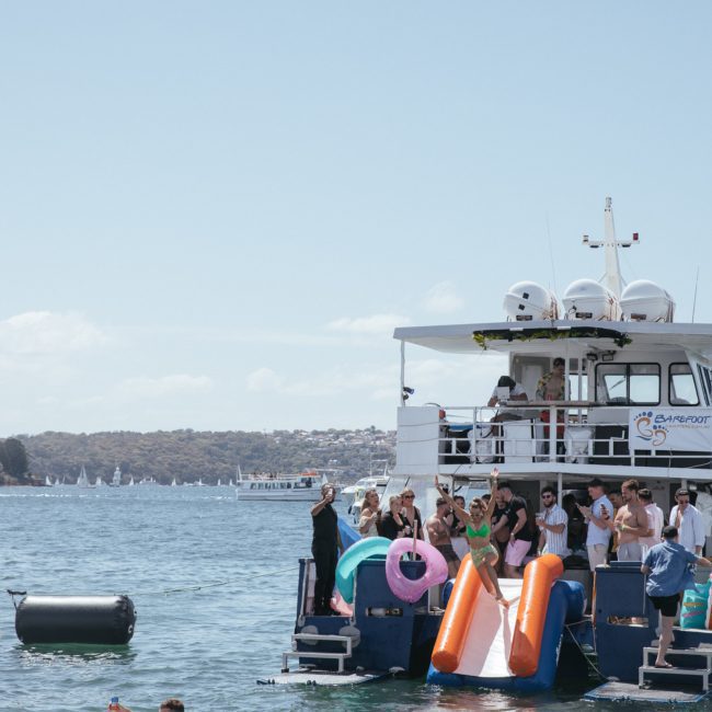 People enjoying a sunny day on and around two boats, with inflatables in the water and an adjacent shoreline in the background. Luxury yacht hire Sydney makes for an unforgettable experience.