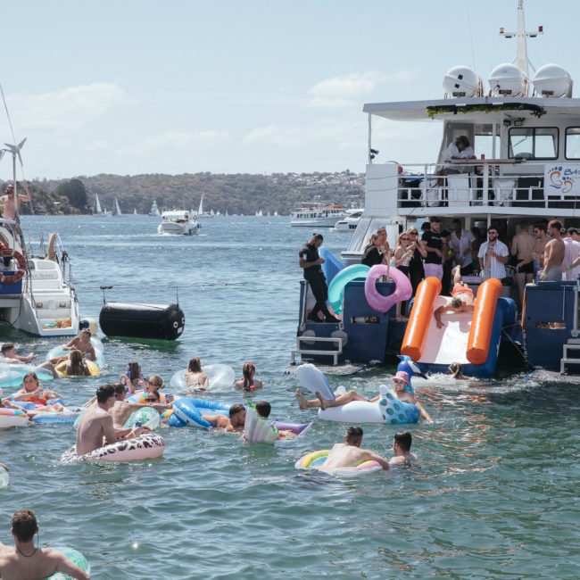 A group of people on a private yacht charter in Sydney Harbour use inflatable slides to enter the lake while others float on inflatable rafts and tubes. Other boats and a picturesque shoreline are visible in the background.