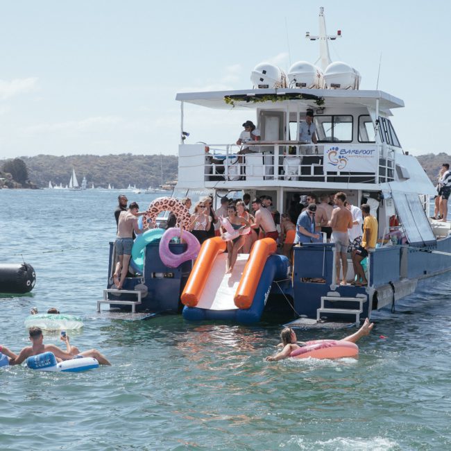 A group of people enjoy a sunny day on a boat, equipped with an inflatable slide, while others float nearby in the water using inflatable tubes and rafts. Ideal for Sydney boat party hire or corporate boat events Sydney, it's perfect for making unforgettable memories on the water.