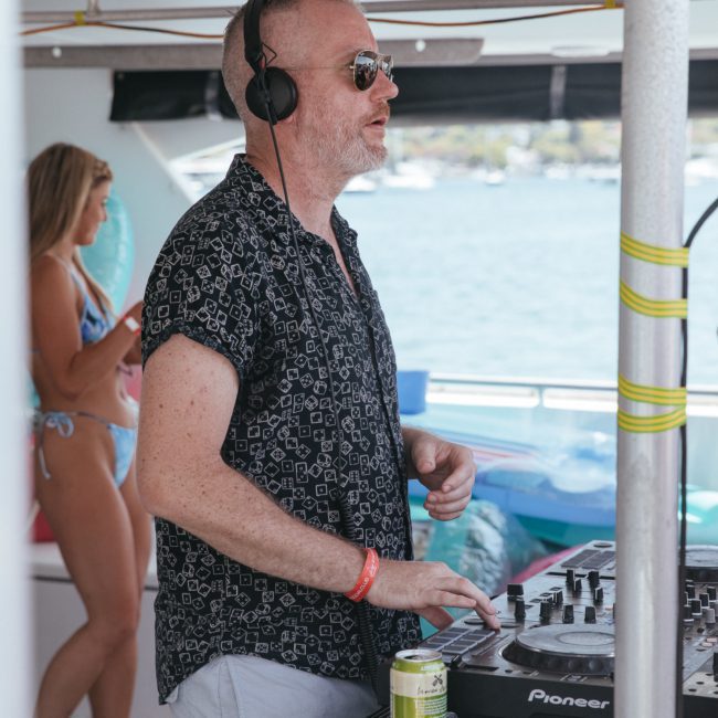 A person wearing headphones operates a DJ controller on a boat. A beverage can is on the table nearby, and a person in swimwear stands in the background. Perfect for a lively Sydney boat party hire or an exclusive DJ boat hire Sydney experience.