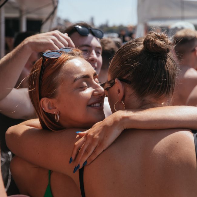 Two women embrace and smile at an outdoor event, surrounded by a crowd of people in swimwear under a tent on a sunny day, enjoying the lively atmosphere of a Sydney boat party hire.