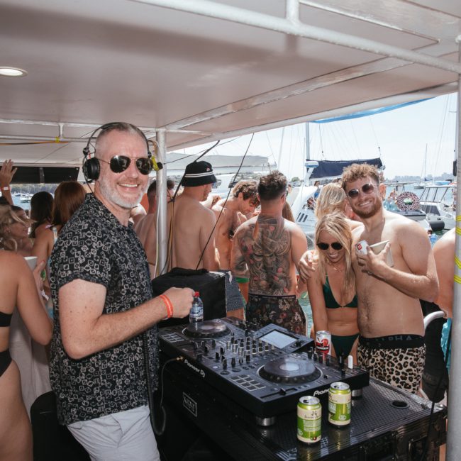 People enjoying music on a boat party. A person in sunglasses and headphones is at the DJ booth from a DJ boat hire Sydney. Others are dancing and mingling around. Cans of drinks are visible on a table.