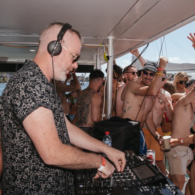 A DJ wearing headphones and a patterned shirt performs on a boat while a crowd of people dance and interact around him. Some attendees are holding drinks and dressed in swimwear, enjoying the Sydney boat party hire on the private yacht charter Sydney Harbour.