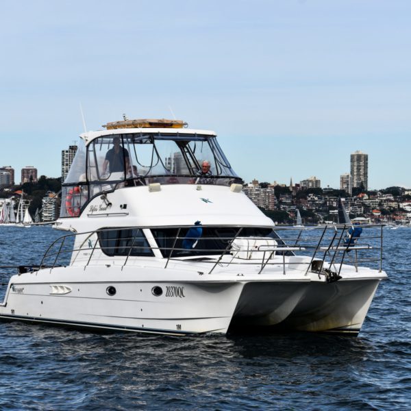 A white catamaran yacht sails on calm water with a city skyline and several smaller boats in the background, perfect for a private yacht charter in Sydney Harbour.