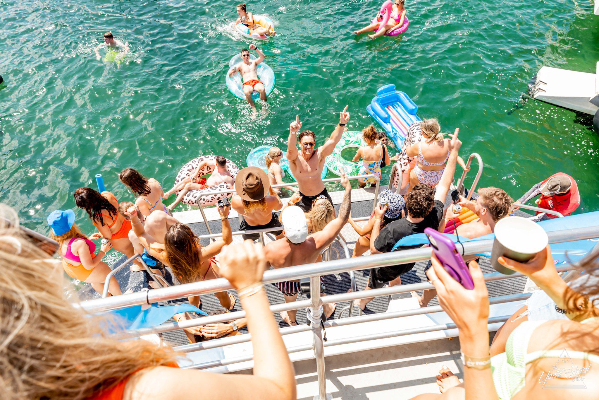 A group of people enjoying a sunny day on a private yacht charter in Sydney Harbour, swimming, lounging on inflatables in the water, and socializing on the deck.