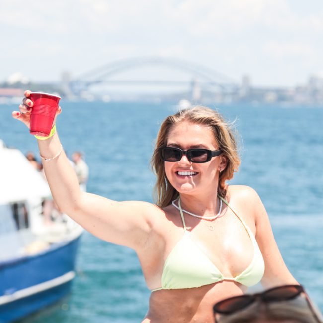 A woman in a green bikini top and sunglasses raises a red cup with a smile on a boat during a catamaran party in Sydney. The Sydney Harbour Bridge is visible in the background.