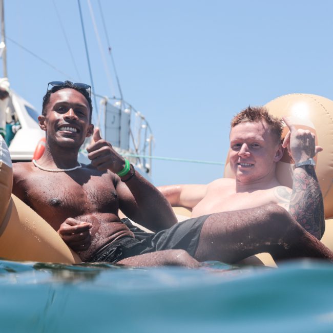 Two men smiling and giving thumbs up while relaxing on an inflatable float in the water, with a luxury yacht hire Sydney and other people in the background.