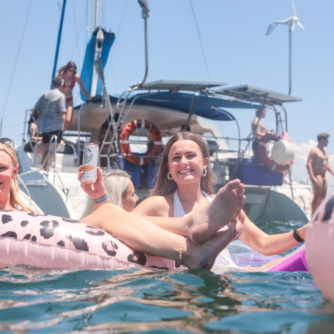 Two smiling women relax on leopard print inflatable floats in the water, holding drinks. In the background, a luxury yacht hire Sydney with people on board glides under a clear, sunny sky.