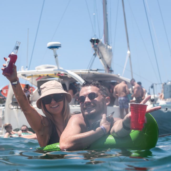 A man and woman enjoy drinks while floating in the water near a luxury yacht. Other people and boats are in the background, creating an ambiance akin to a vibrant DJ boat hire Sydney event.