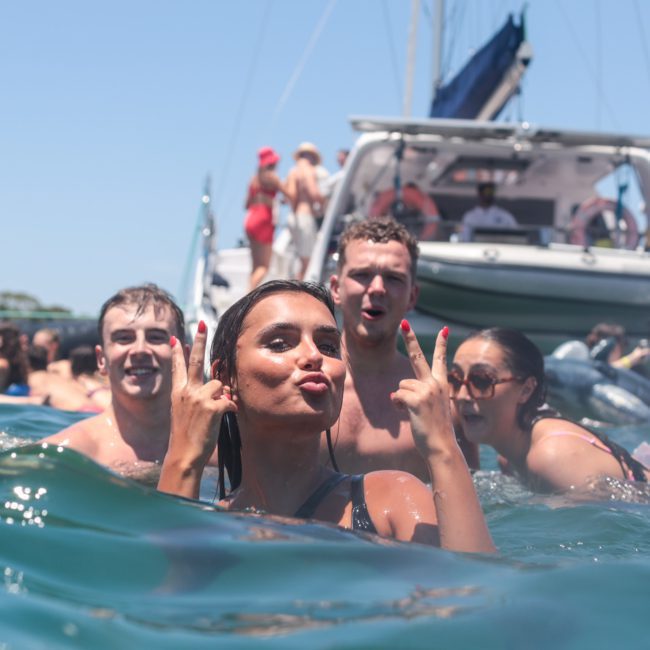 A group of people swim and pose for the camera in the water near a boat. One person in the foreground makes a peace sign with both hands during a lively Sydney boat party hire.