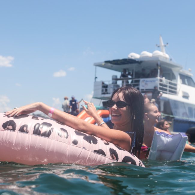 A woman is floating on a leopard print inflatable in the water, with a boat and other people in the background enjoying a lively Sydney boat party hire on a sunny day.