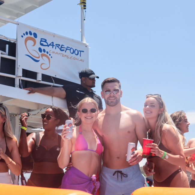 Group of people in swimsuits smiling and holding drinks on a boat with a "Barefoot Charter" sign in the background, perfectly capturing the essence of private yacht charter Sydney Harbour.