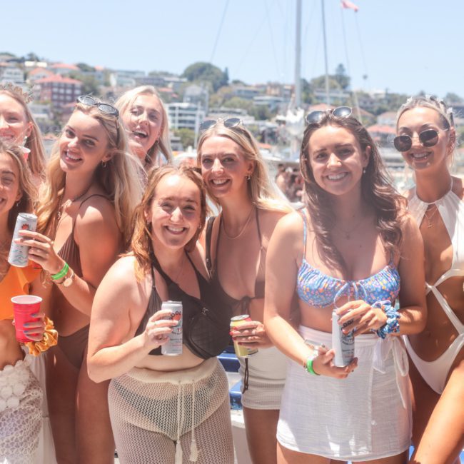 A group of people in swimwear smile for a photo while holding drinks at an outdoor gathering near a body of water with boats, possibly from a luxury yacht hire Sydney, in the background.