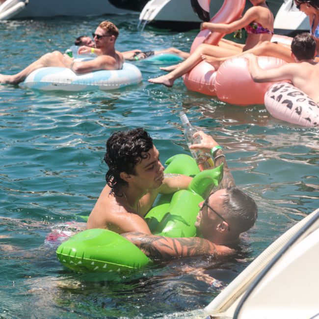 Two people are in the water playing with a green inflatable, while others relax on inflatable floats nearby. Boats, including a private yacht charter on Sydney Harbour, are anchored in the background on a sunny day.