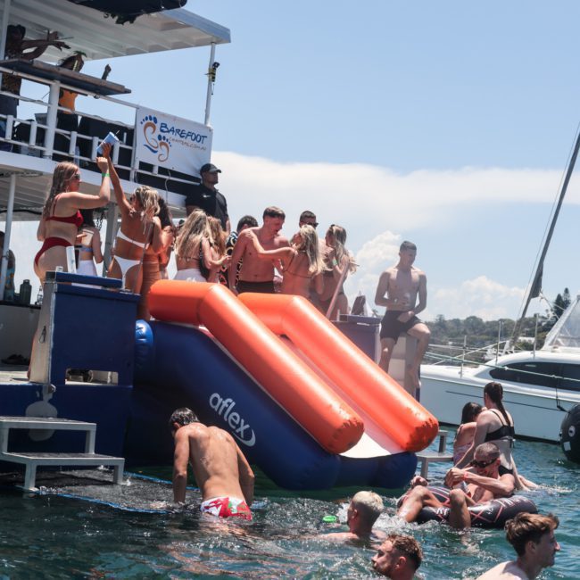 A group of people enjoy water activities, including using red inflatable slides, from a docked boat while others swim nearby in a sunny setting. Several boats are visible in the background, creating a lively atmosphere perfect for a Sydney boat party hire.