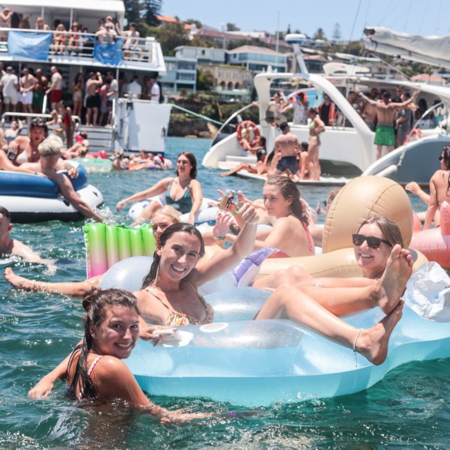 A group of people enjoying a sunny day on a body of water. Many are using various inflatables while boats and yachts are docked nearby. Some people are smiling and waving at the camera during a lively Sydney boat party hire or private yacht charter in Sydney Harbour.