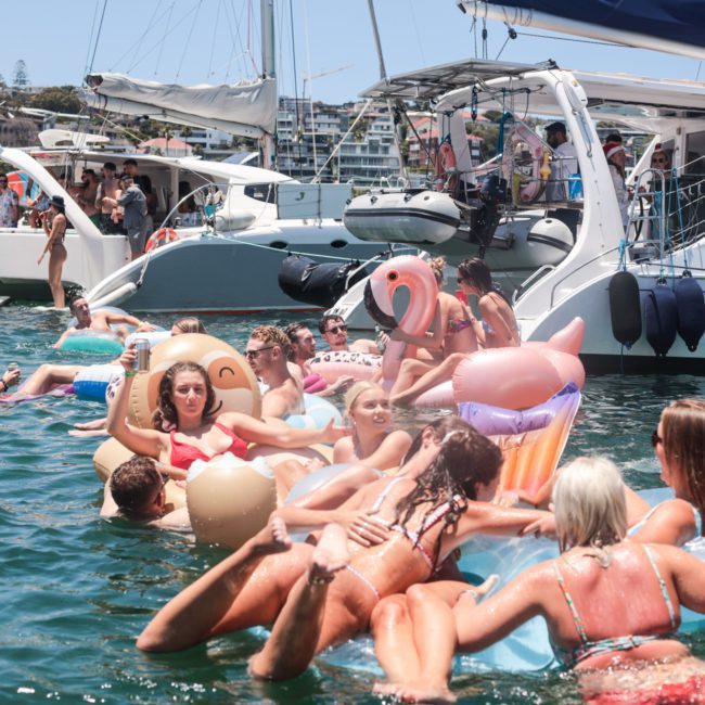 People on inflatable rafts and floaties relax in the water near docked boats on a sunny day, enjoying a luxury yacht hire Sydney.