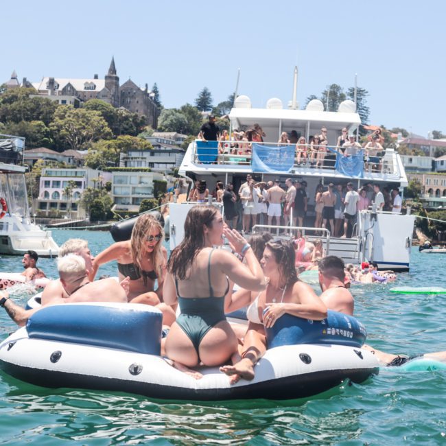 A group of people enjoys a sunny day on inflatable rafts and boats in a bustling waterfront area with buildings and trees in the background, possibly considering a private yacht charter Sydney Harbour for their next adventure.