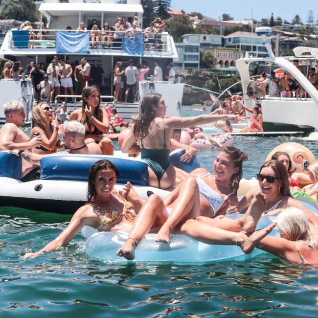 Group of people enjoying a summer day on the water, with some on inflatables and others socializing on boats in the background. Consider Sydney boat party hire for your next event to make your day as vibrant and enjoyable.