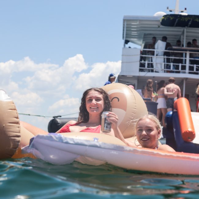 Two people smile while floating on an inflatable in the water. A boat with a slide and several people on board is in the background, showcasing the fun of a Sydney boat party hire.